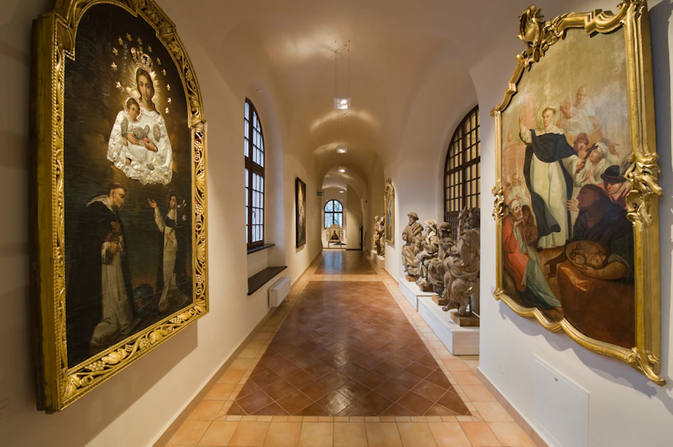 A museum corridor with large religious paintings, statues, and arched windows under a vaulted ceiling.