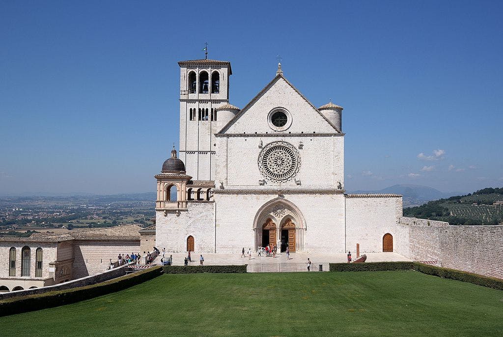 Basilique historique en pierre blanche avec une grande rosace, un clocher et une pelouse bien entretenue. Des personnes marchent près de l'entrée.