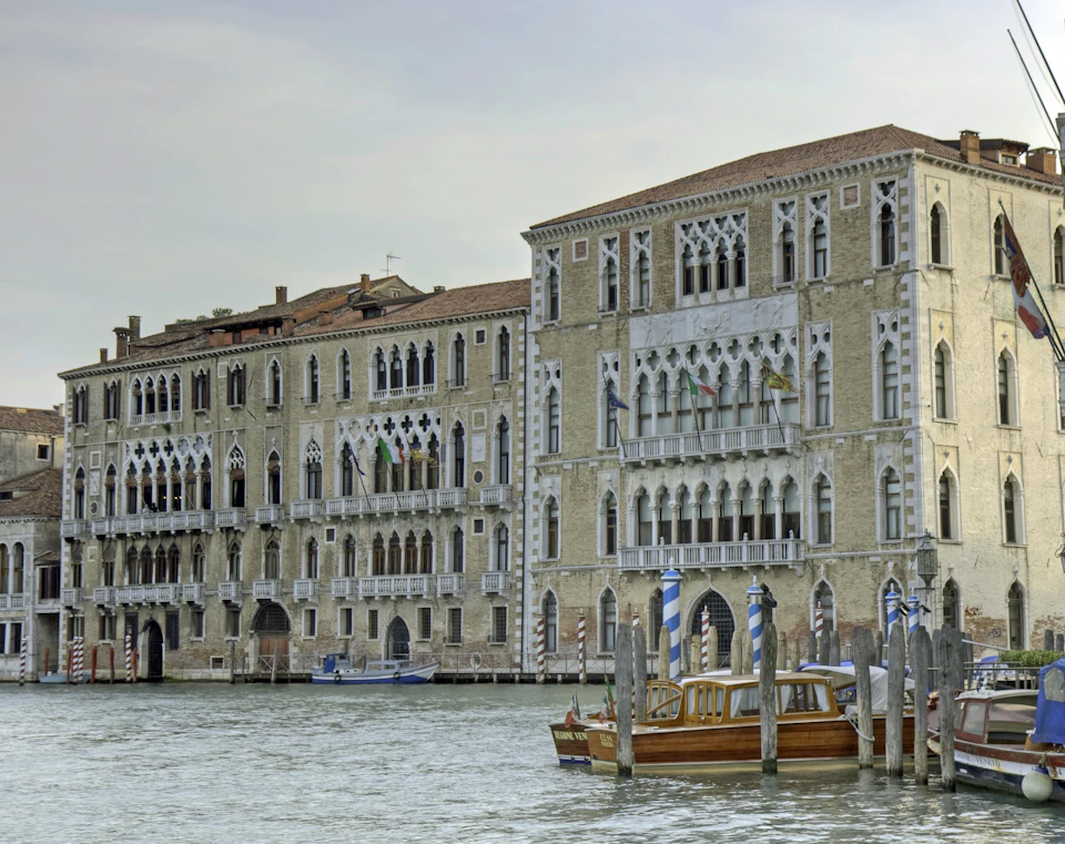 Two historic buildings with arched windows along a canal, several boats docked in the foreground.
