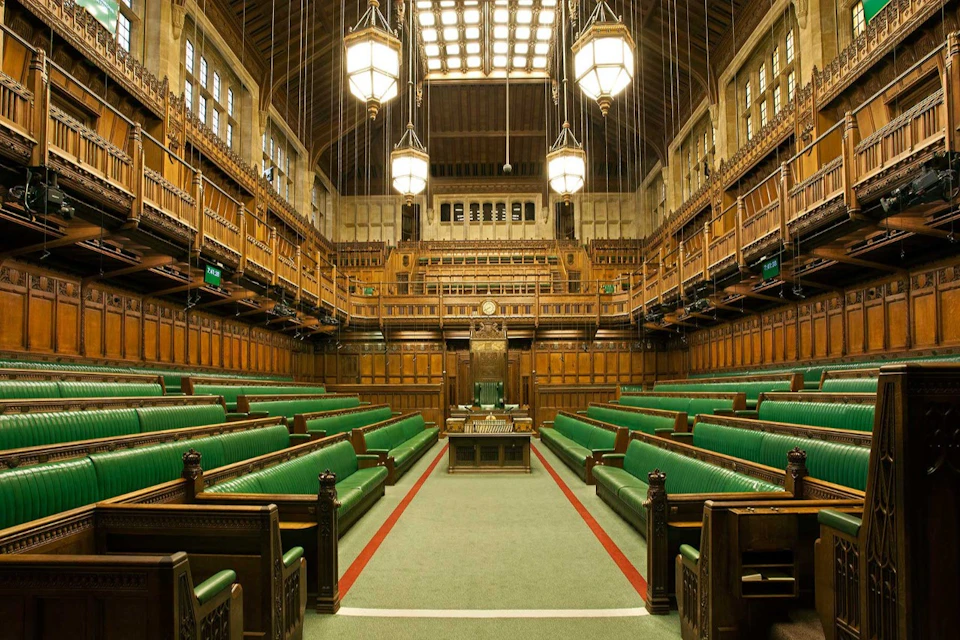 A legislative chamber with green benches, wooden paneling, a central speaker's chair, and hanging chandeliers.