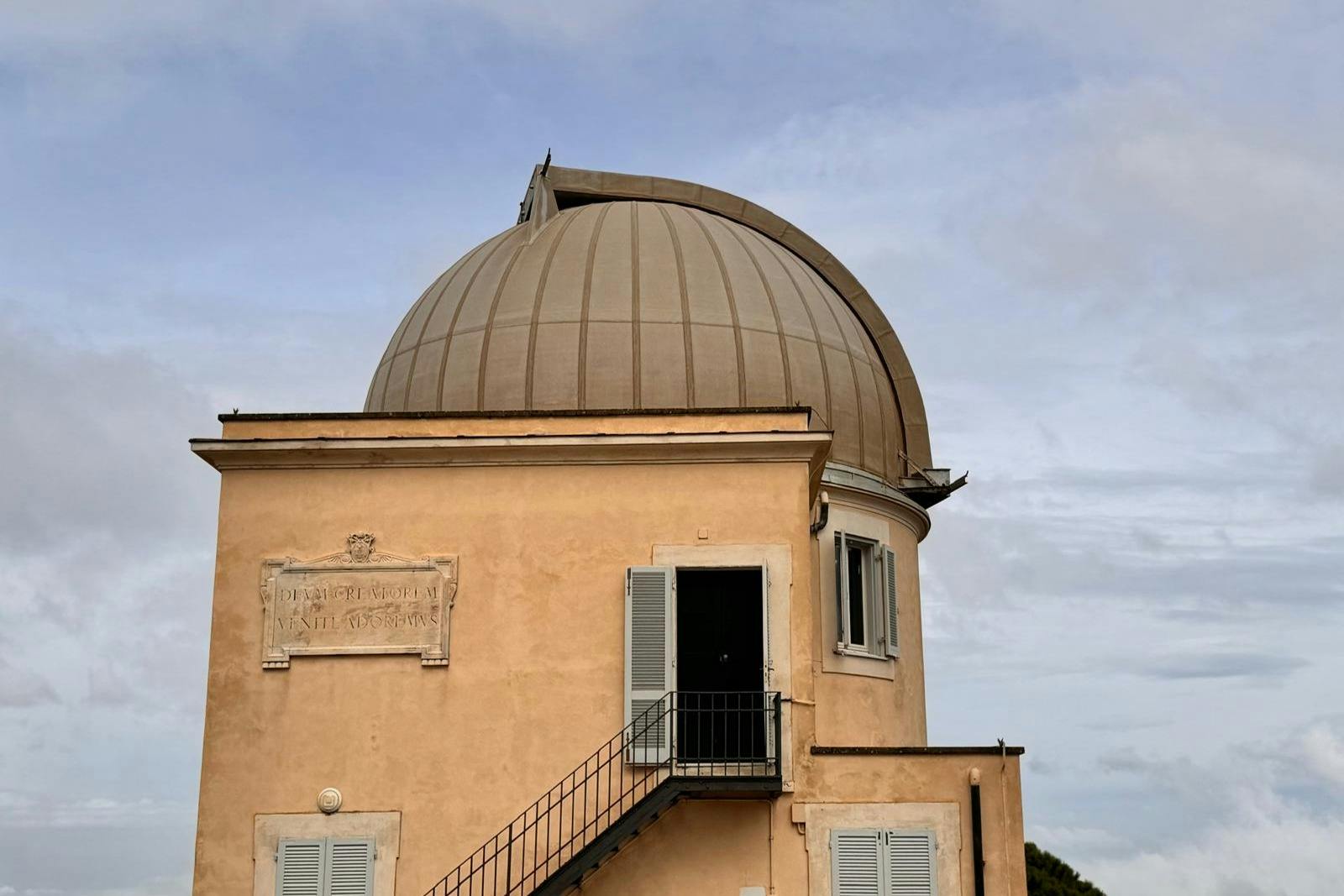 Building with a large, dome-shaped roof, two shuttered windows, an open door with a metal staircase, and a plaque on the wall.