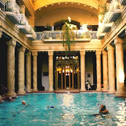 En inomhuspool med människor som simmar, omgiven av utsmyckade pelare, med balkong och hängande växter ovanför.