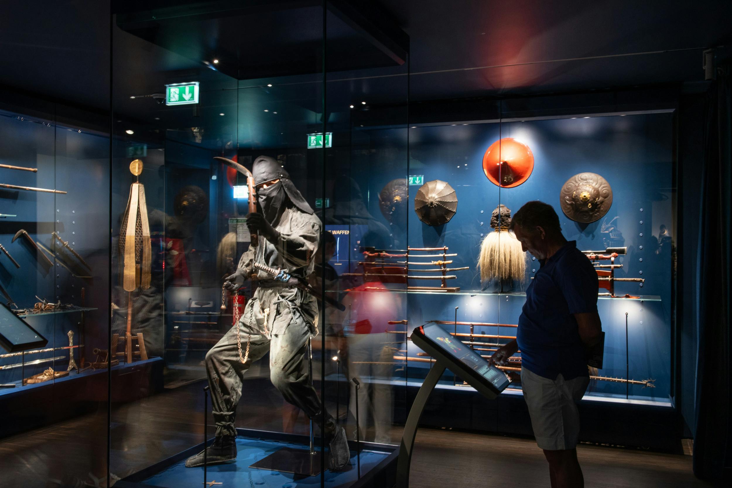 Una exposició de museu amb una figura de ninja en una caixa de vidre, amb armes i escuts a la paret del darrere.