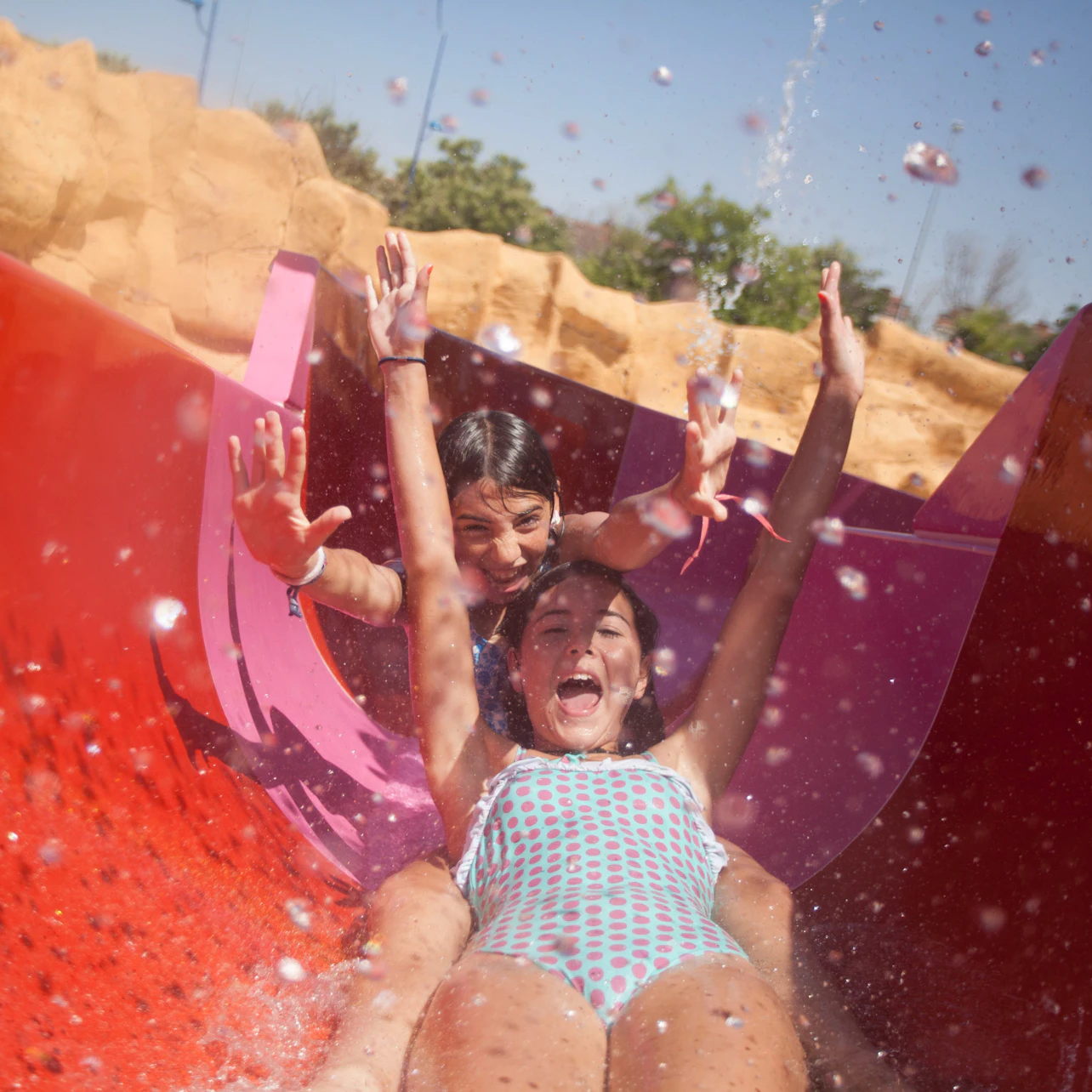 Two people slide down a colorful waterslide, splashing water around them, with a sunny outdoor backdrop.