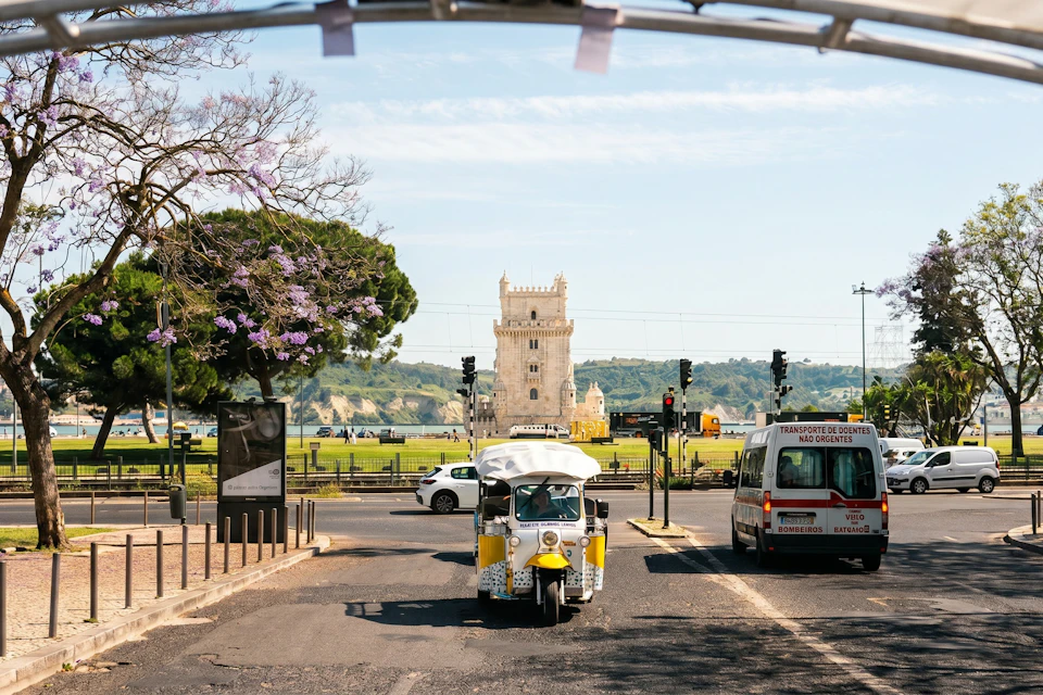 A tuk-tuk and a van at a crossroad with the Torre de Belém in the background on a sunny day.