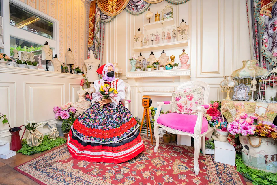 A person in traditional attire sits in a decorated room with floral arrangements, a pink chair, and various ornaments.