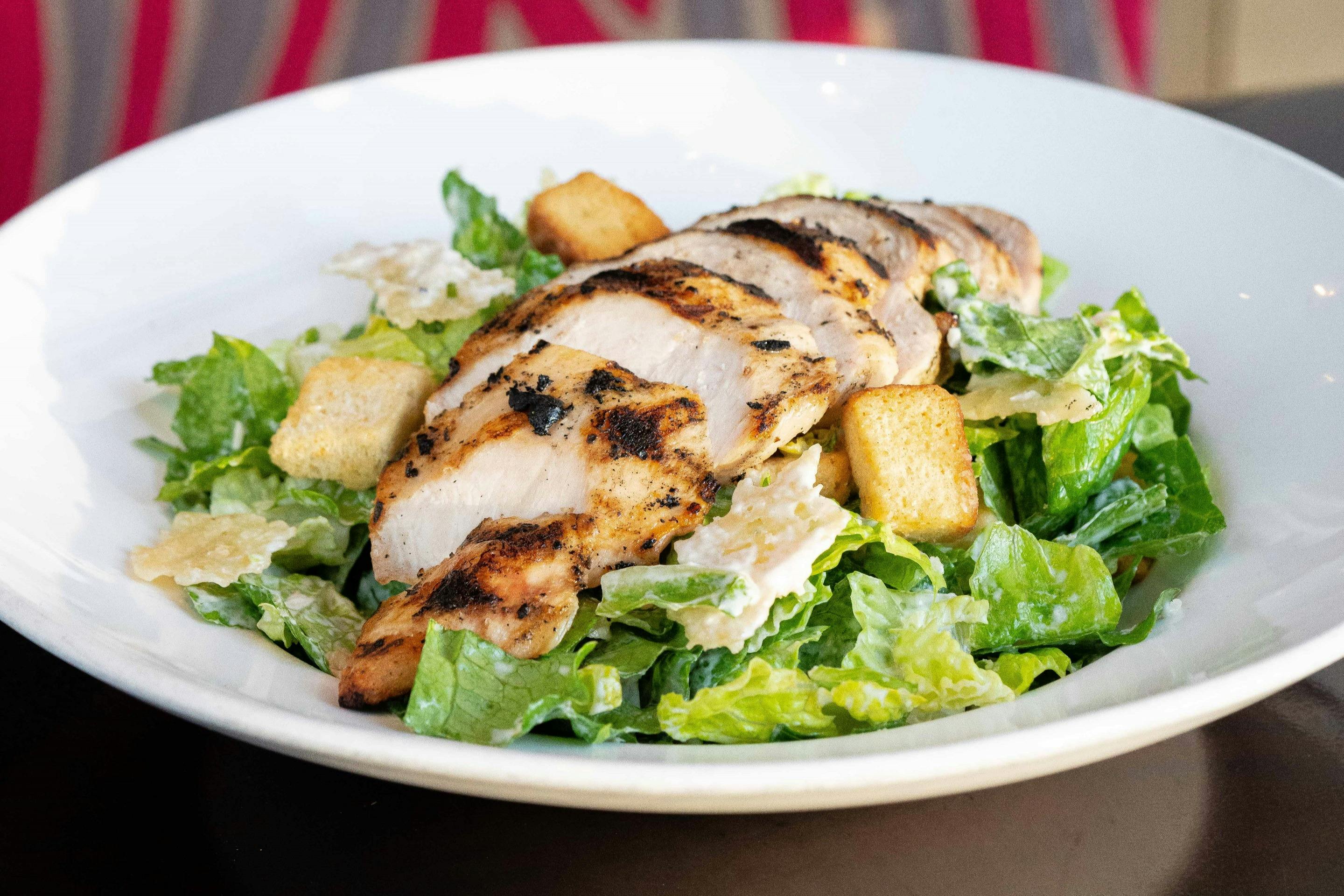 흰색 접시에 구운 치킨 슬라이스, 로메인 상추, 크루통, 파르메산 치즈를 곁들인 시저 샐러드.