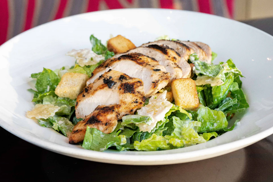 A Caesar salad with grilled chicken slices, romaine lettuce, croutons, and Parmesan cheese on a white plate.