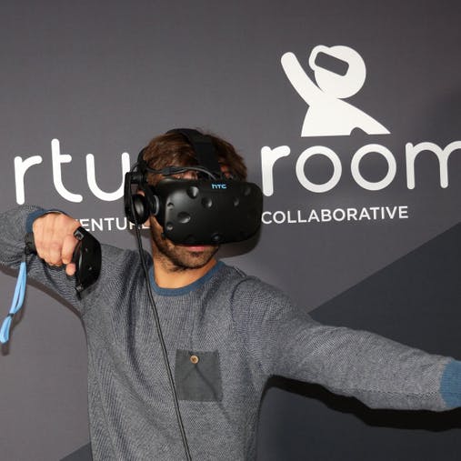 A person wearing a VR headset and holding controllers, standing in front of a wall with the text "Virtual Room."