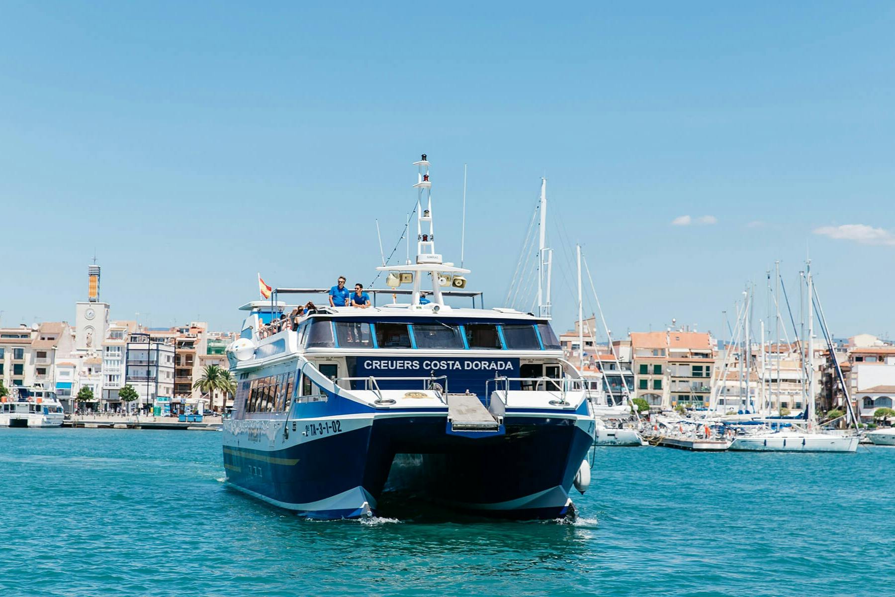 Um ferry chamado "Creuers Costa Dorada" a navegar em águas azuis perto de um porto com edifícios e veleiros em fundo.
