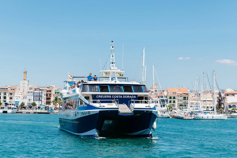 A ferry named 'Creuers Costa Dorada' sailing on blue water near a harbor with buildings and sailboats in the background.