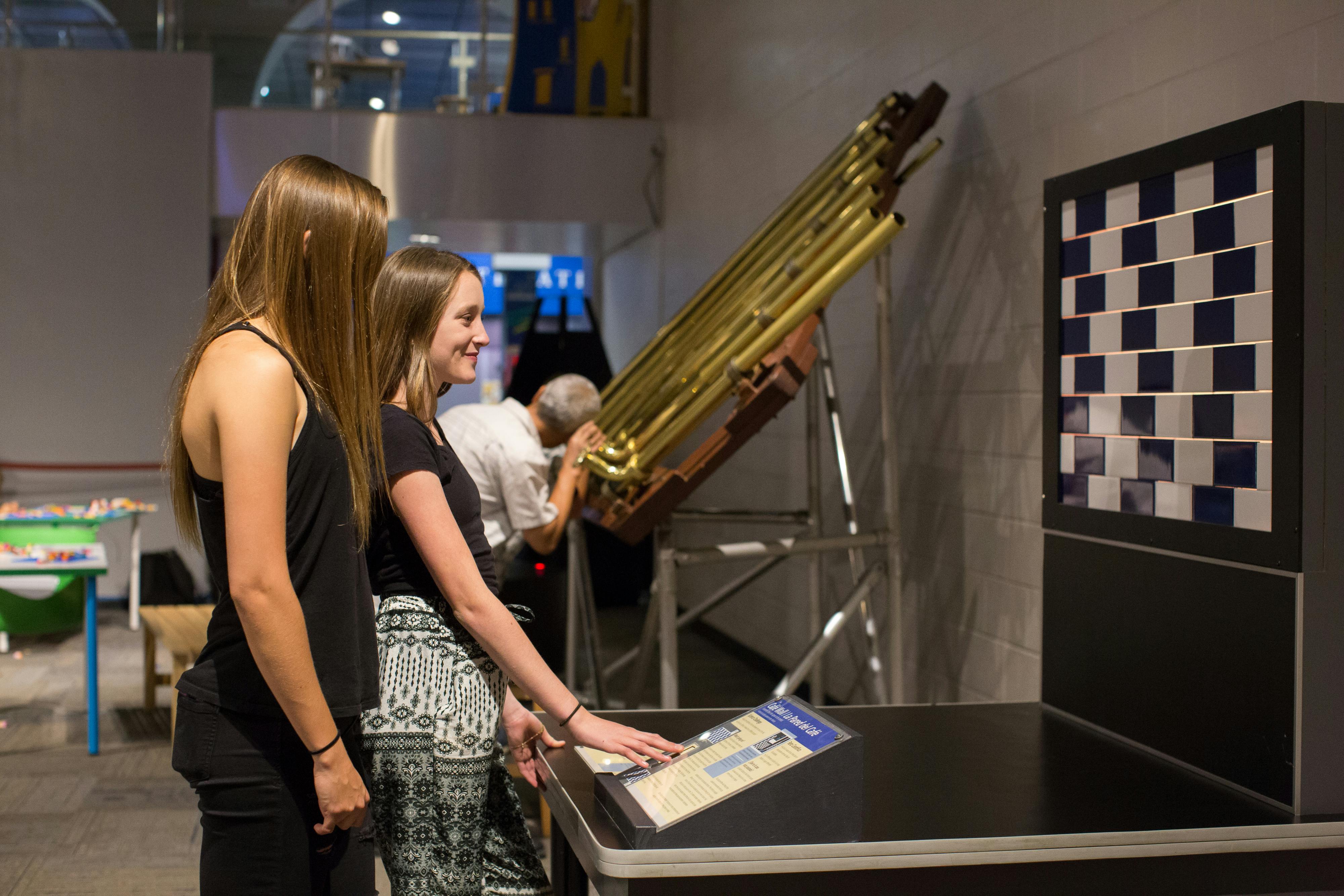 Δύο νεαρές γυναίκες αλληλεπιδρούν με ένα έκθεμα που διαθέτει ορειχάλκινους σωλήνες και έναν πίνακα ελέγχου. Ένας ηλικιωμένος άνδρας παρατηρεί στο παρασκήνιο.