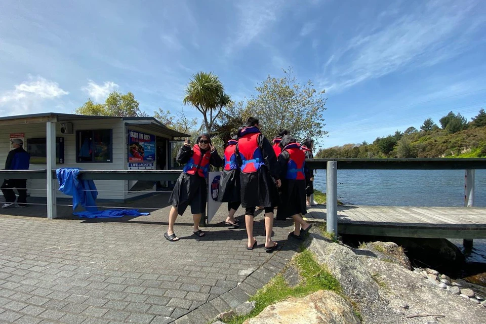 A group of people wearing red and blue life jackets standing on a dock near a building and a body of water.