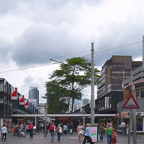 Busy urban street with people walking, various shops including a Burger King, and modern buildings under a cloudy sky.