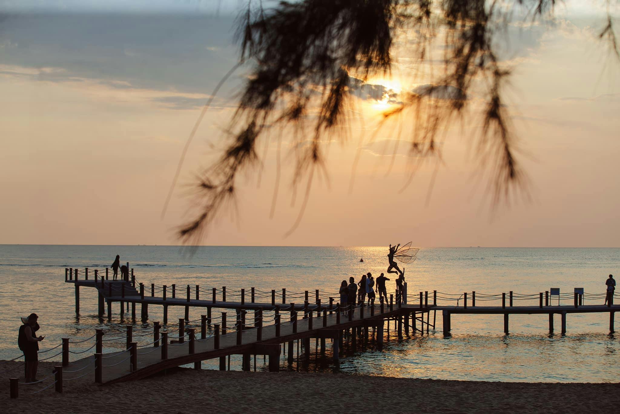 A pier over the ocean at sunset with people gathered near a sculpture, framed by tree branches.