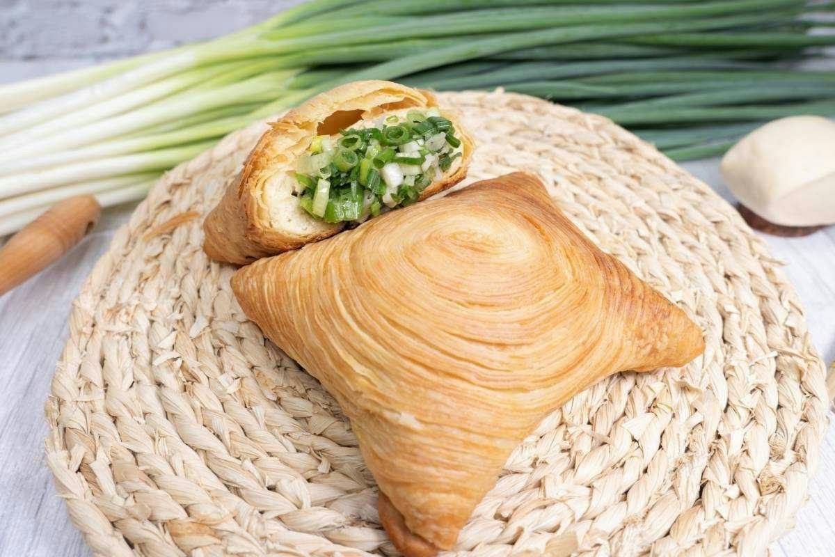 A golden, flaky pastry on a woven mat, with one piece cut open showing a filling of green onions.