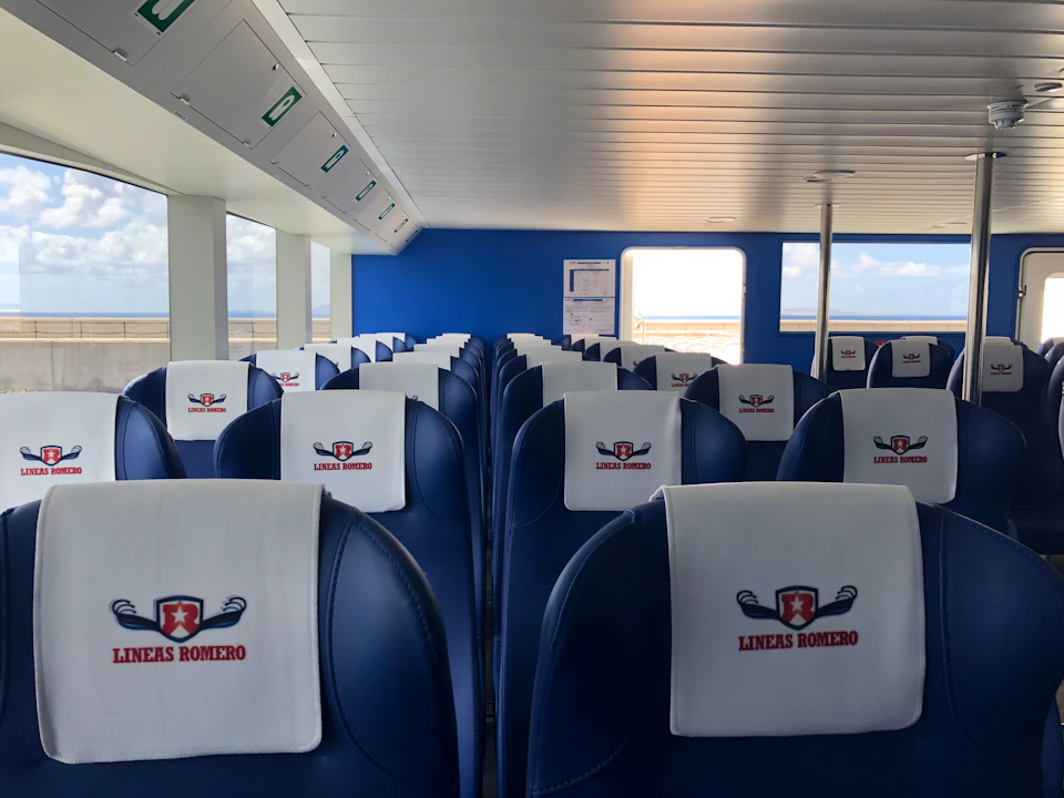 Interior of a ferry with rows of navy blue seats, each with a white cover displaying the "LINEAS ROMERO" logo.