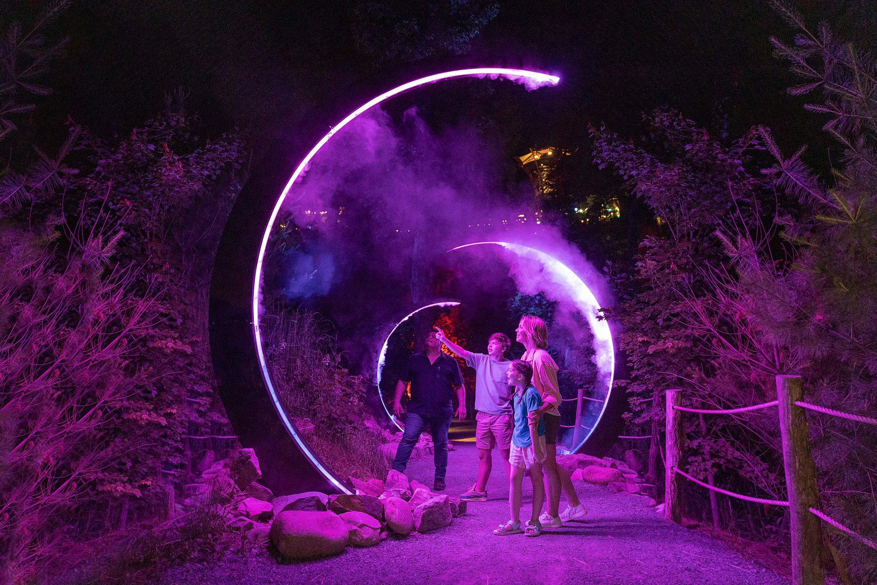 People walk through illuminated circular arches with purple lighting at night, surrounded by trees and foliage.