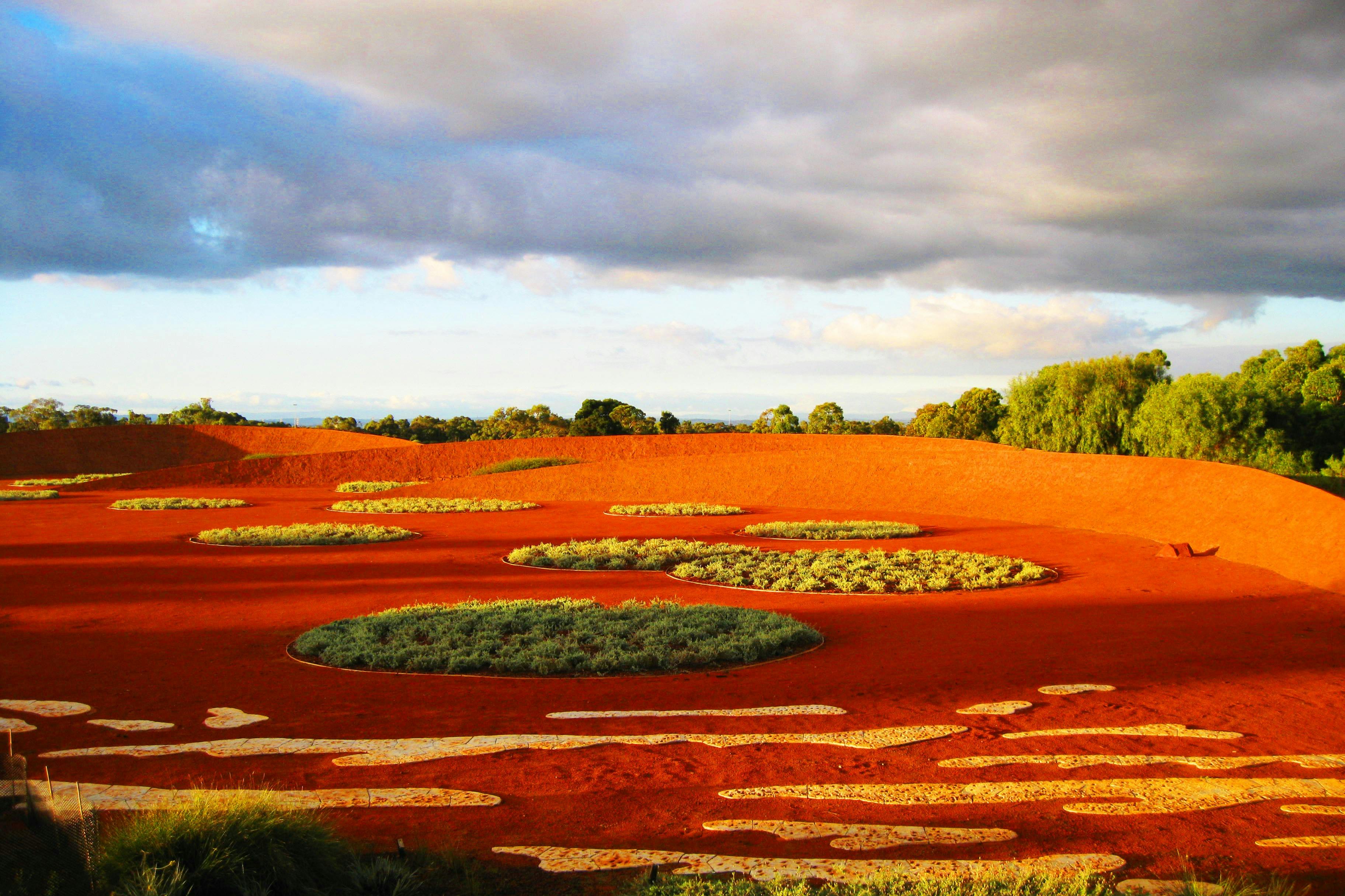 Živá červená krajina s kulatými skvrnami zelené vegetace, obklopená stromy a pod zataženou modrou oblohou.