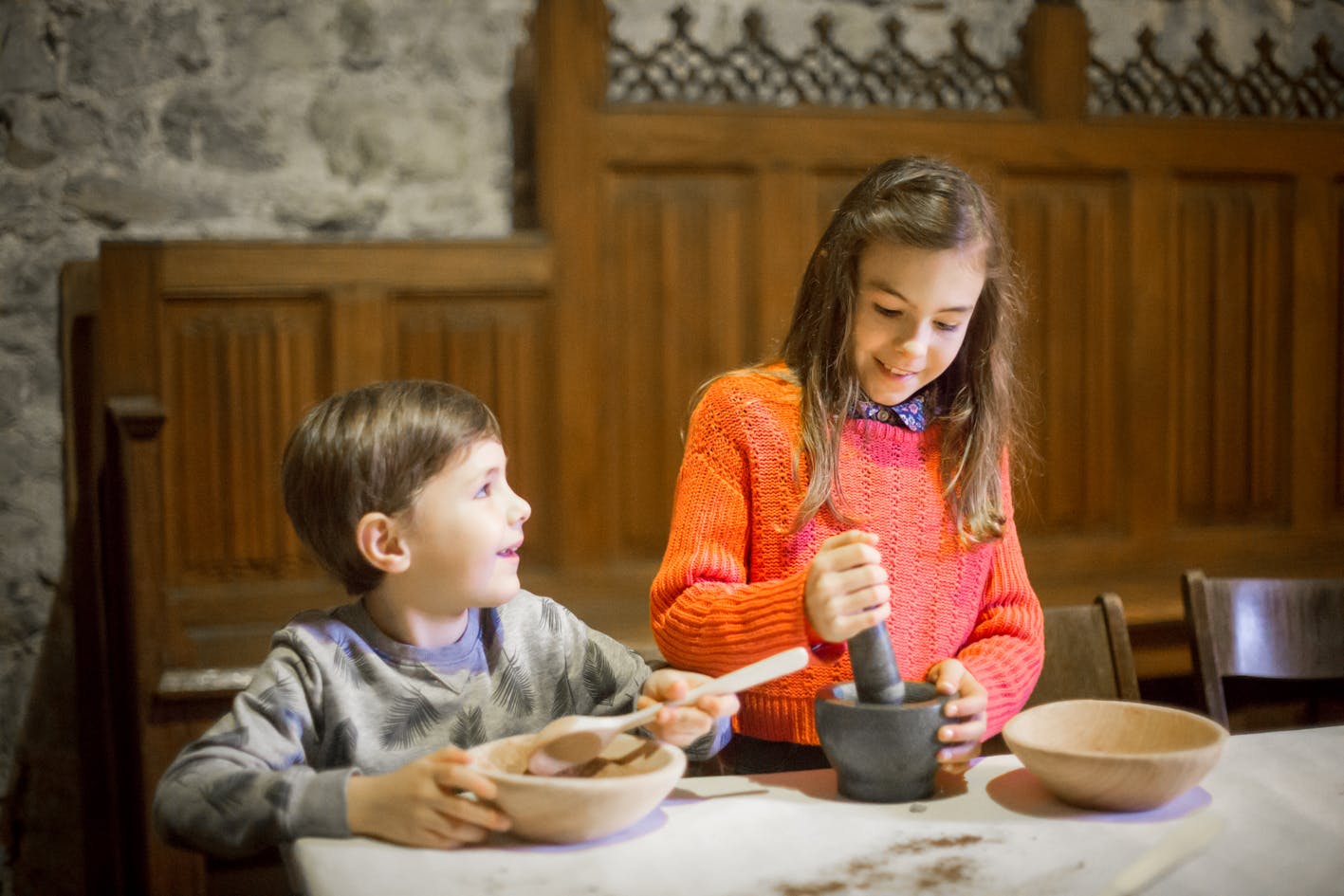 A boy and a girl sit at a table with wooden bowls; the girl uses a mortar and pestle while the boy observes.