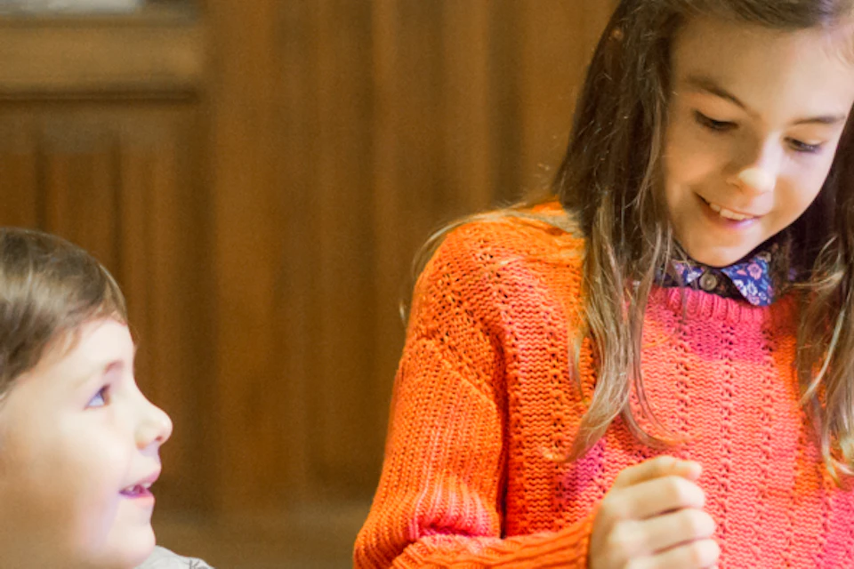 A boy and a girl sit at a table with wooden bowls; the girl uses a mortar and pestle while the boy observes.