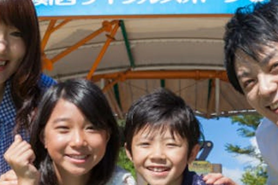 A smiling family of four poses outdoors in front of a sign that reads "関西サイクルスポーツセンター" in Japanese.
