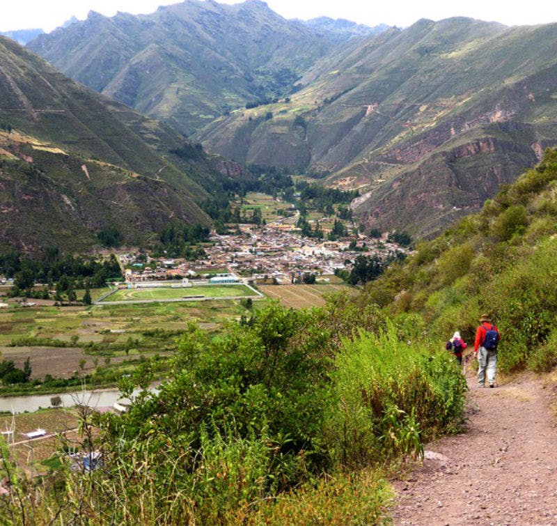 Hikers walking along a mountain trail overlooking a valley and small town, surrounded by green hills and distant peaks.