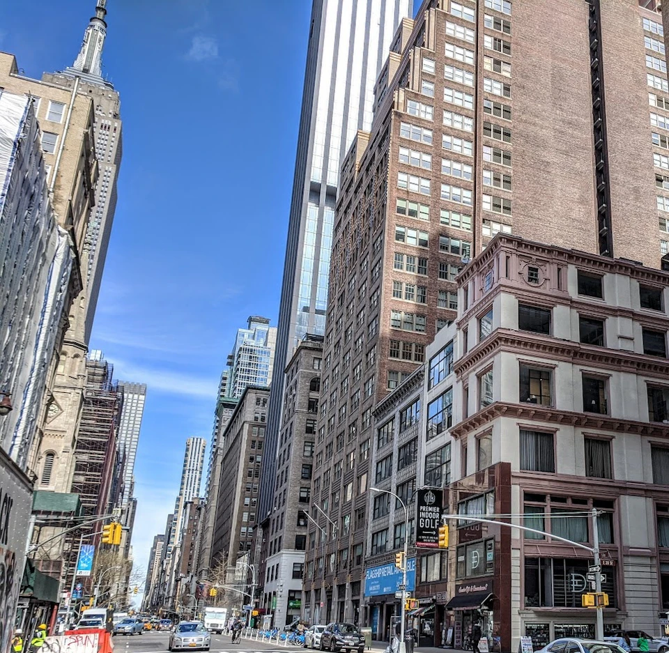 A city street with tall buildings under a blue sky, featuring cars, pedestrians, and traffic lights in an urban setting.