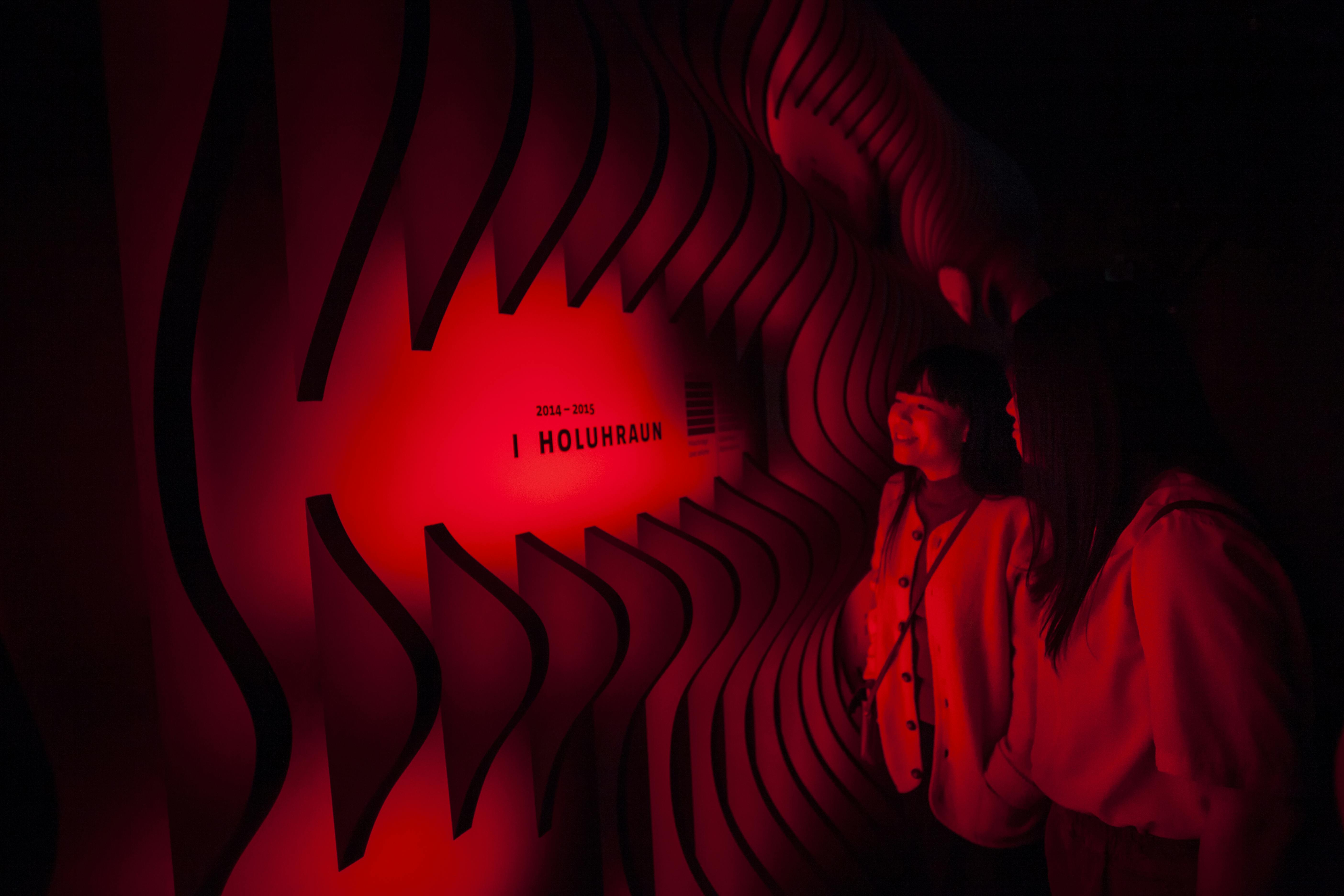 두 사람이 "2014 - 2015 I HOLUHRAUN" 이라는 문구가 새겨진 물결 모양의 붉은 불빛 벽을 바라보고 있습니다.