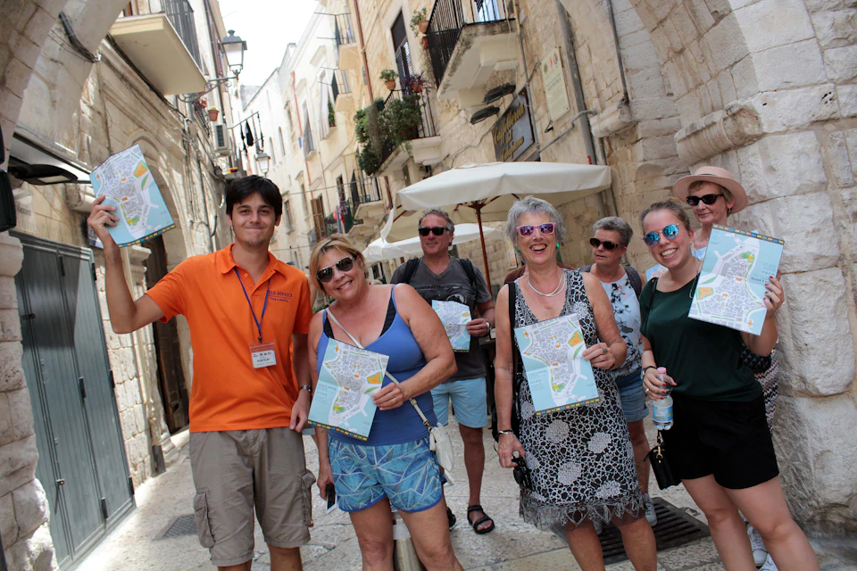 A tour guide holding a map leads three tourists holding maps through a narrow, sunlit street with historic buildings.
