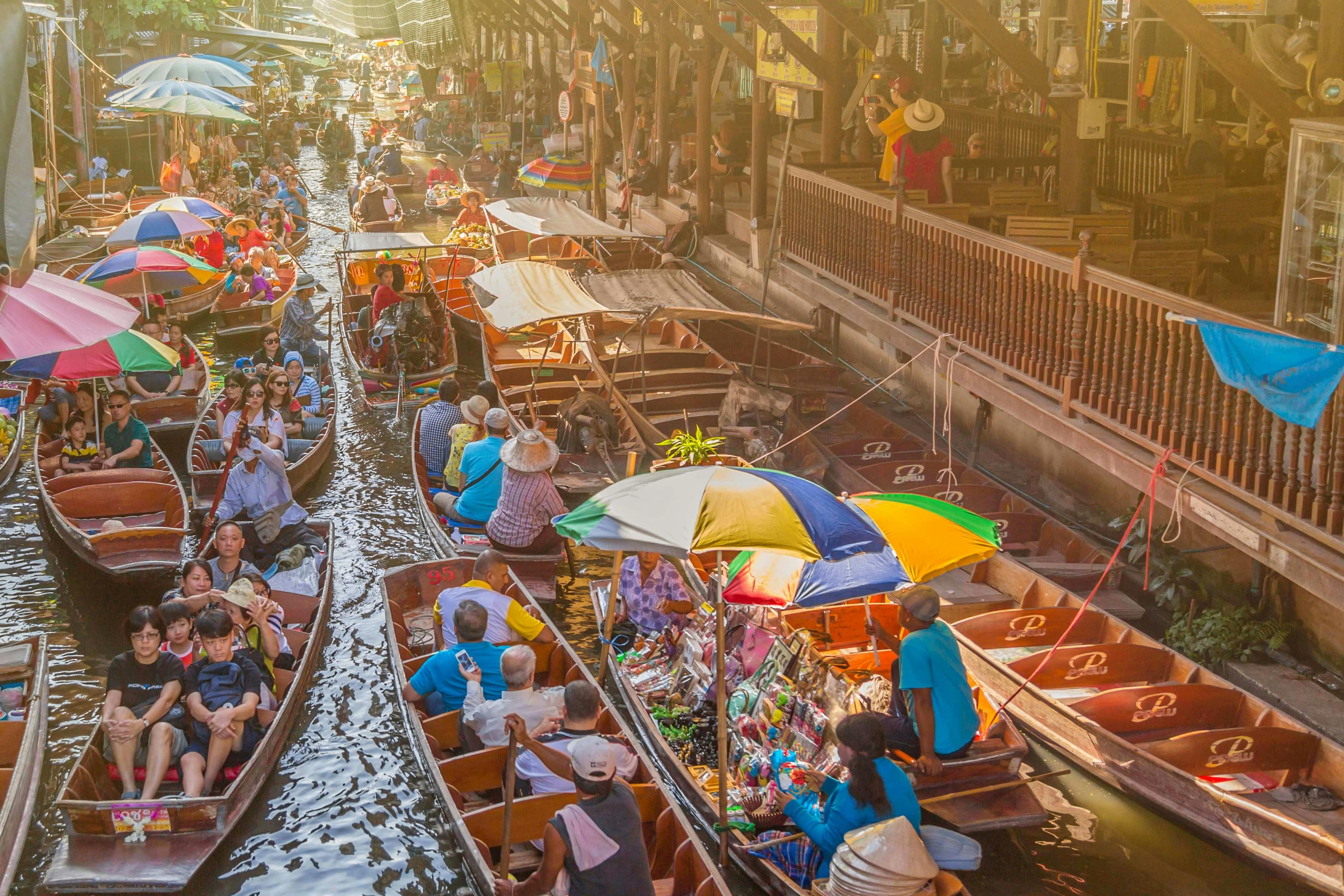 A bustling floating market with numerous boats carrying goods and people under colorful umbrellas on a narrow waterway.