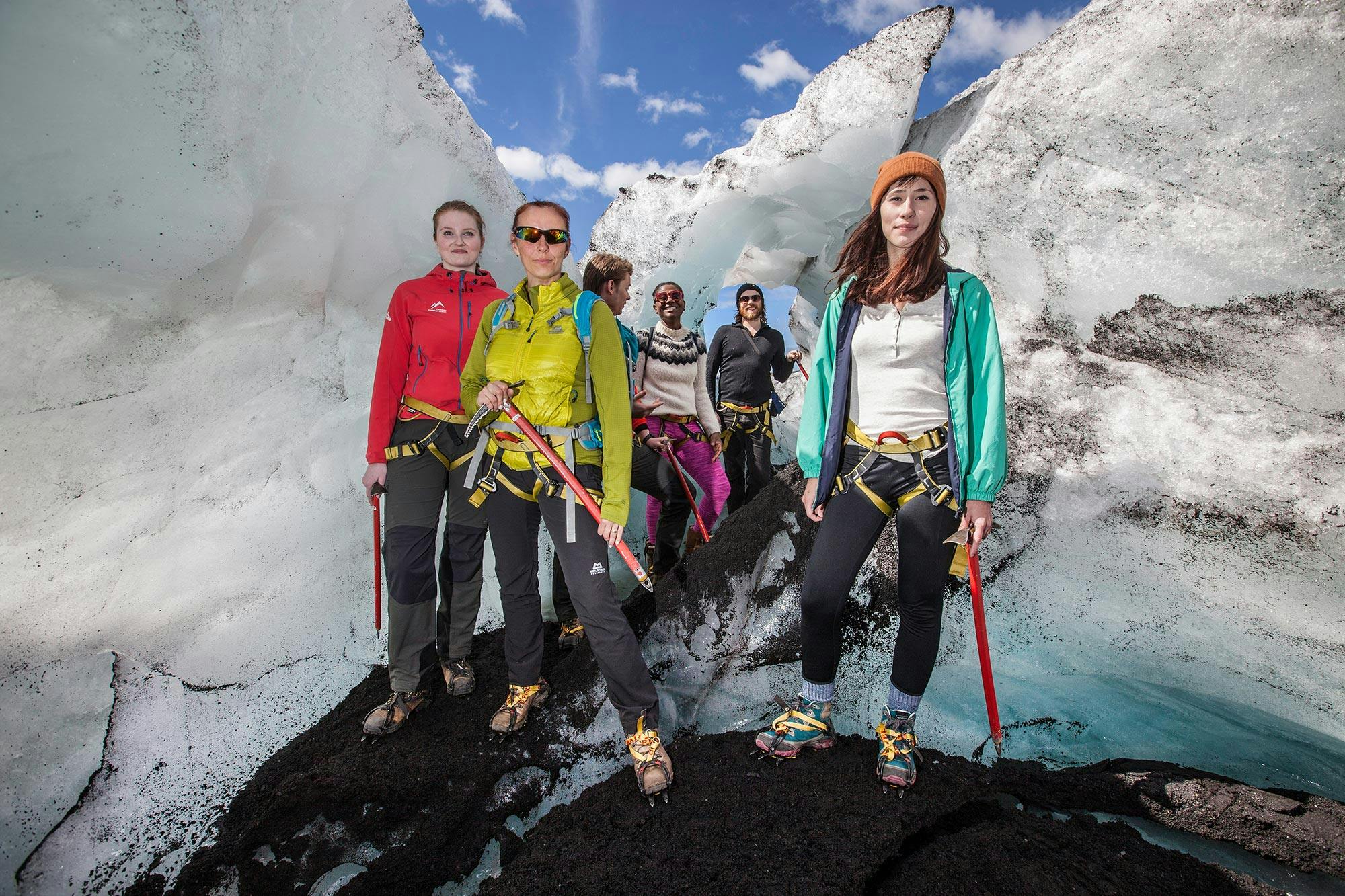 People in climbing gear stand in an ice cave, surrounded by ice walls and wearing crampons on their boots. Bright, sunny day.