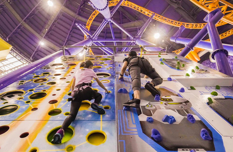 Two people in climbing gear scale a colorful indoor rock climbing wall, with a high ceiling and vivid structures in the background.