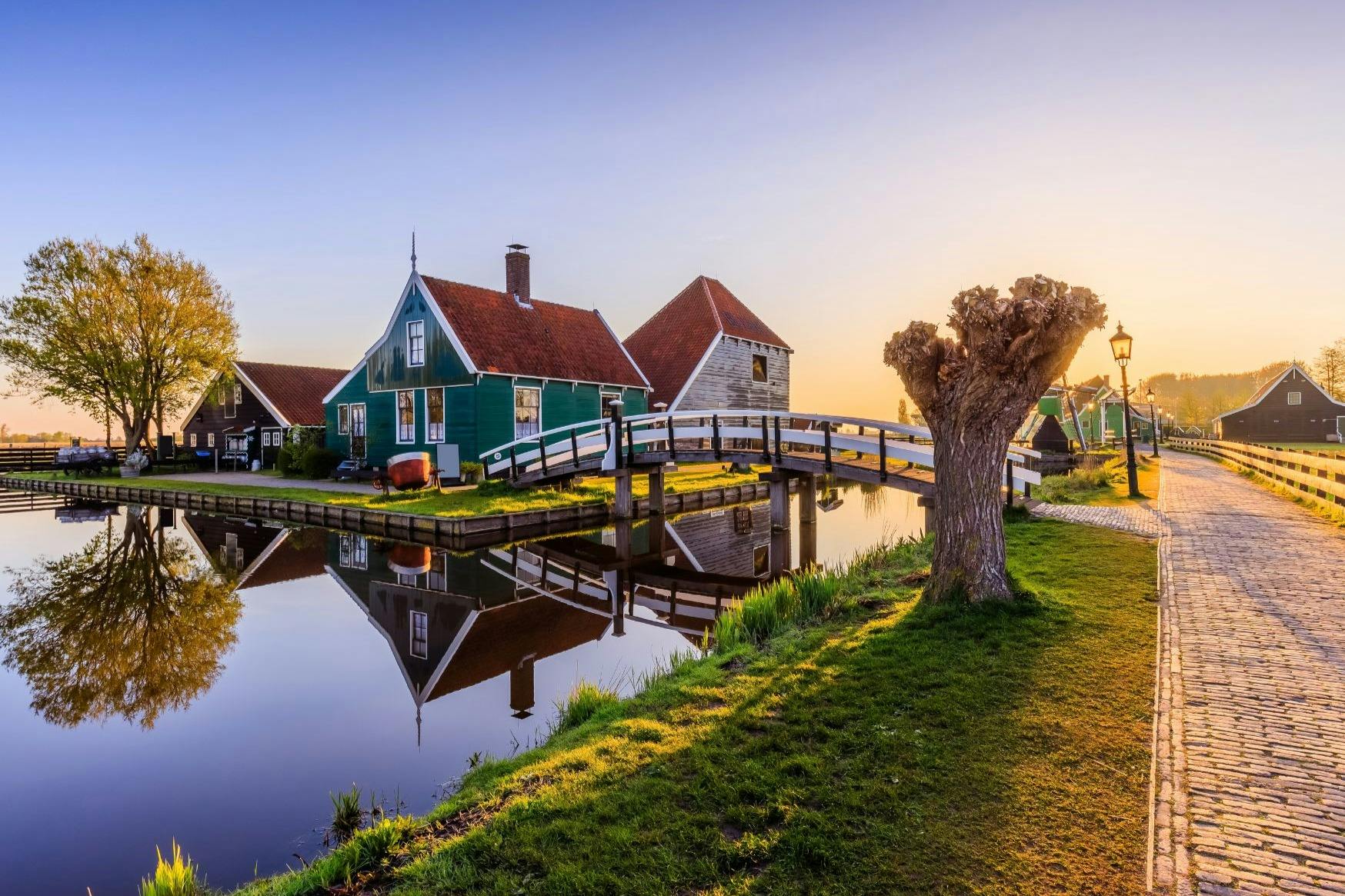 赤い屋根の緑の家、運河の上の歩道橋、夕暮れ時の草原の裸の木。