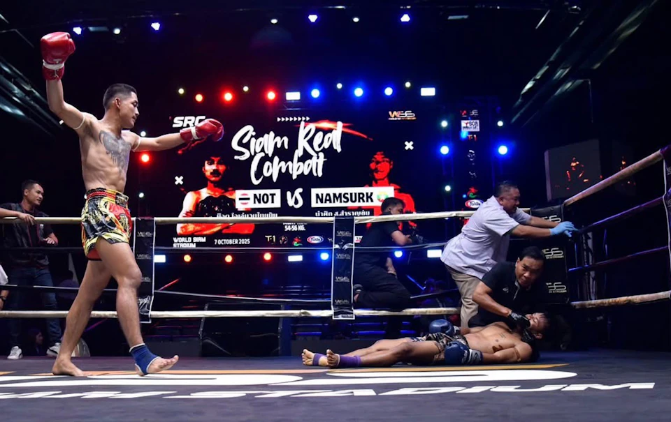 A Muay Thai fighter celebrates in a ring after a match. A defeated opponent lies on the ground. A scoreboard is visible.