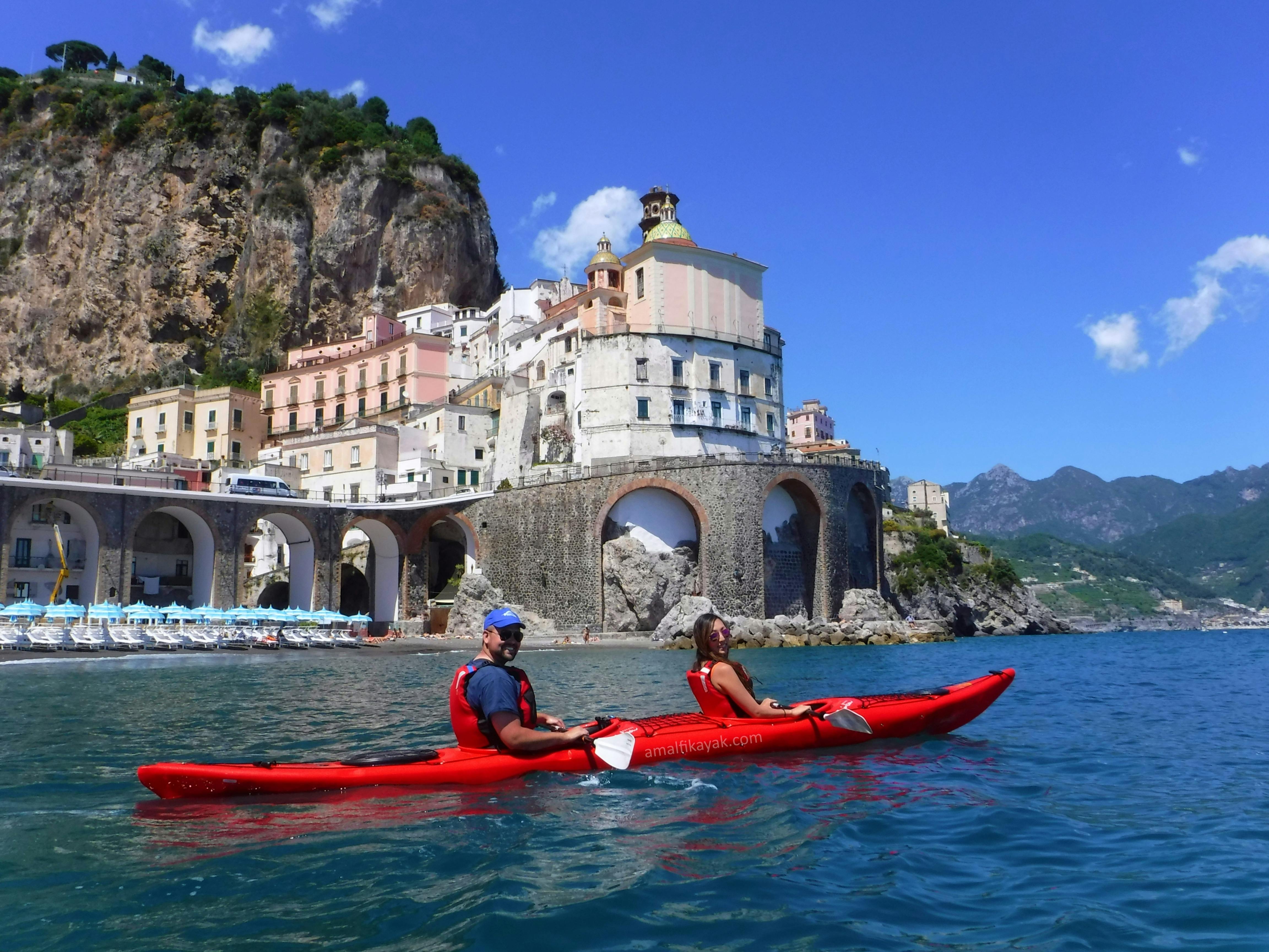 Due persone in kayak rosso remano vicino a una città costiera con grandi archi, edifici colorati e una scogliera sotto un cielo azzurro e limpido.