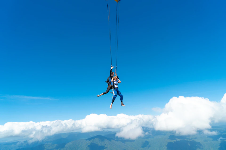 Two people paragliding high above clouds with mountains visible below, under a clear blue sky.