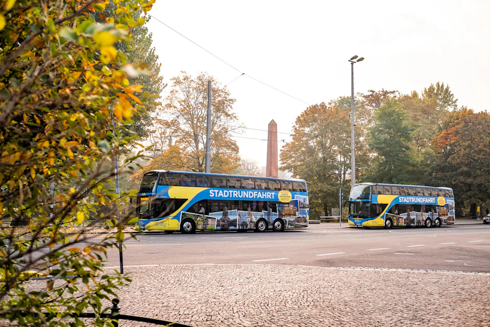 Two double-decker tour buses with "STADTRUNDFAHRT" on the side, parked on a tree-lined street with an obelisk in the background.