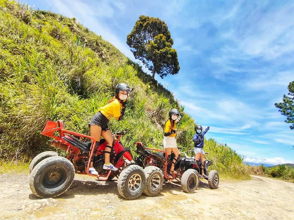 Three people on ATVs parked on a dirt path with greenery and a tree in the background under a bright blue sky.