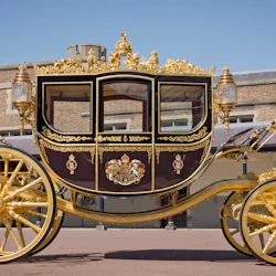 En förgylld, utsmyckad hästdragen vagn med invecklade dekorationer, stora gyllene hjul och kungliga emblem framför en historisk byggnad.