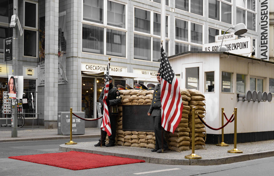 Two guards stand at a checkpoint with sandbags and U.S. flags, in front of a building with "Checkpoint Charlie" signage.