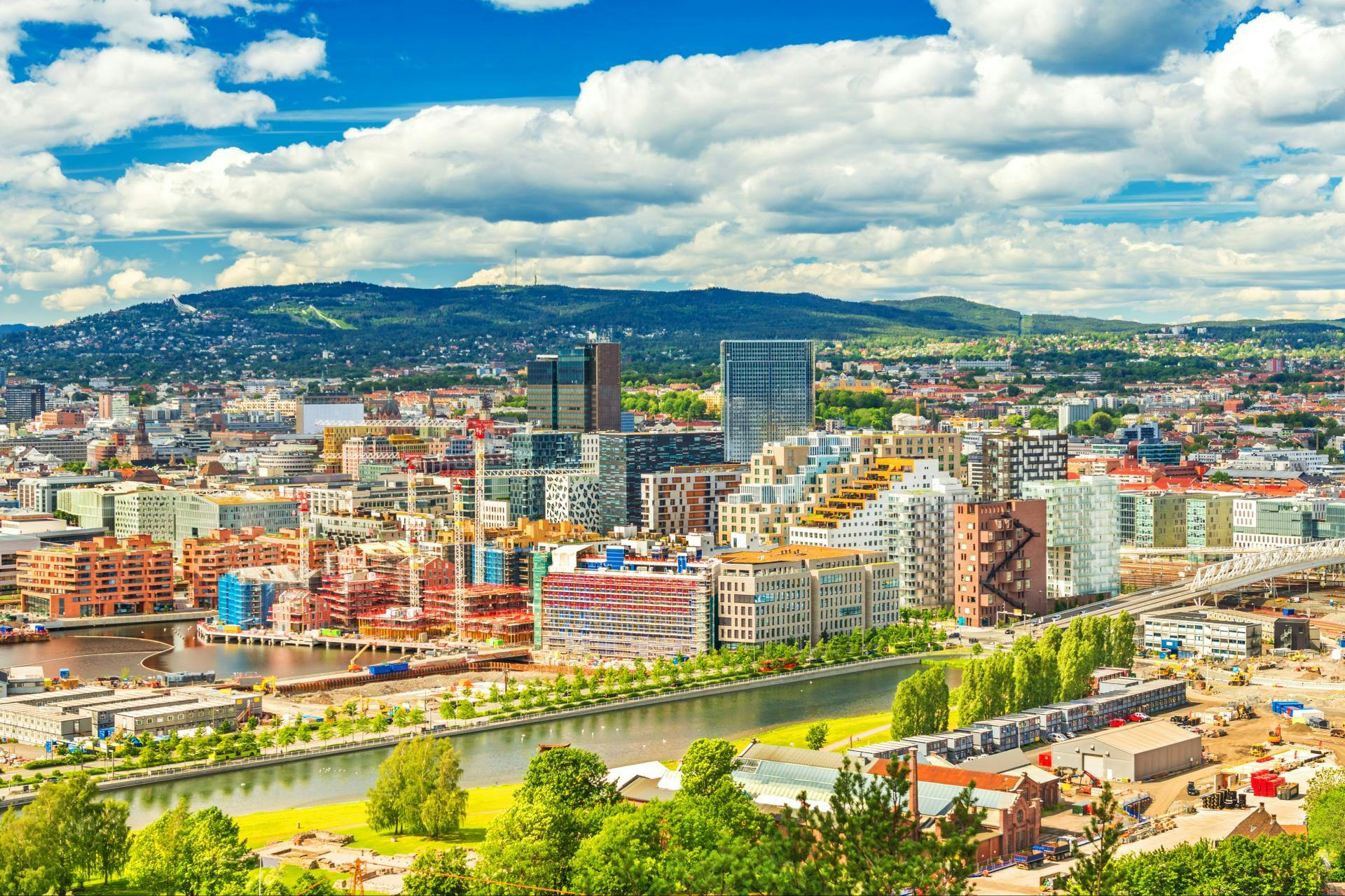 Färgglad stadsbild med moderna byggnader, en flod och omgivande grönska under en delvis molnig himmel med avlägsna kullar.