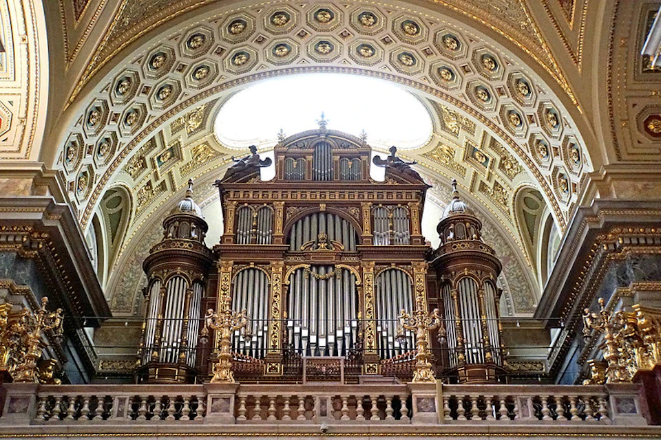 A grand, ornate pipe organ with intricate carvings and gold accents sits under an arched, decorative ceiling in a grand room.