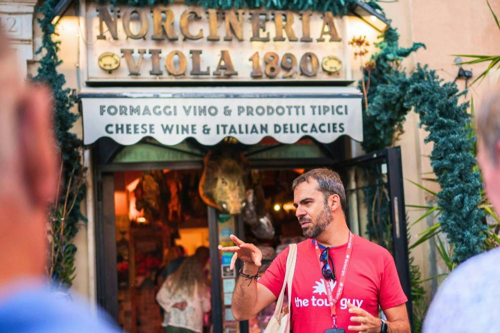 A man in a red shirt gesturing in front of a deli shop with an Italian sign, surrounded by green decorations.
