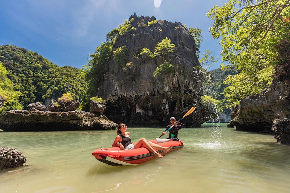 Two people on an inflatable raft in clear water, surrounded by tall, green-clad cliffs under a bright blue sky.