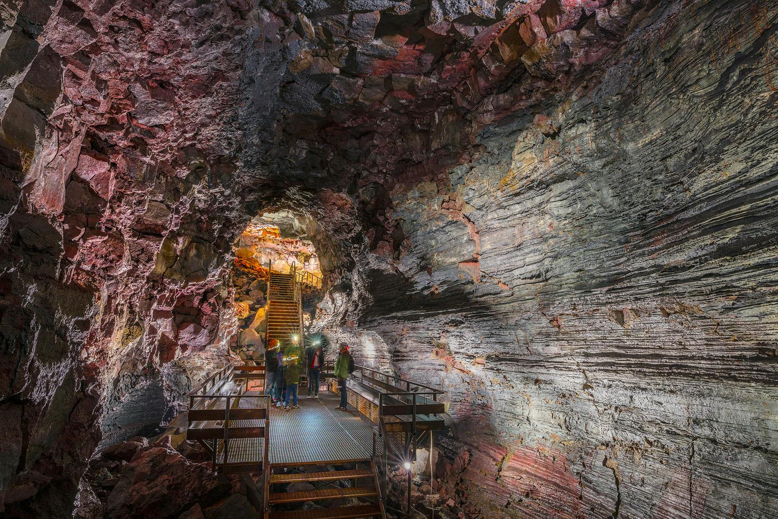 Tourists wearing helmets explore an illuminated volcanic cave with metal walkways and stairs.