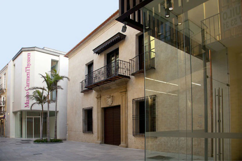 Modern building with glass facade next to an older building with a balcony and iron railings; palm trees line the walkway.