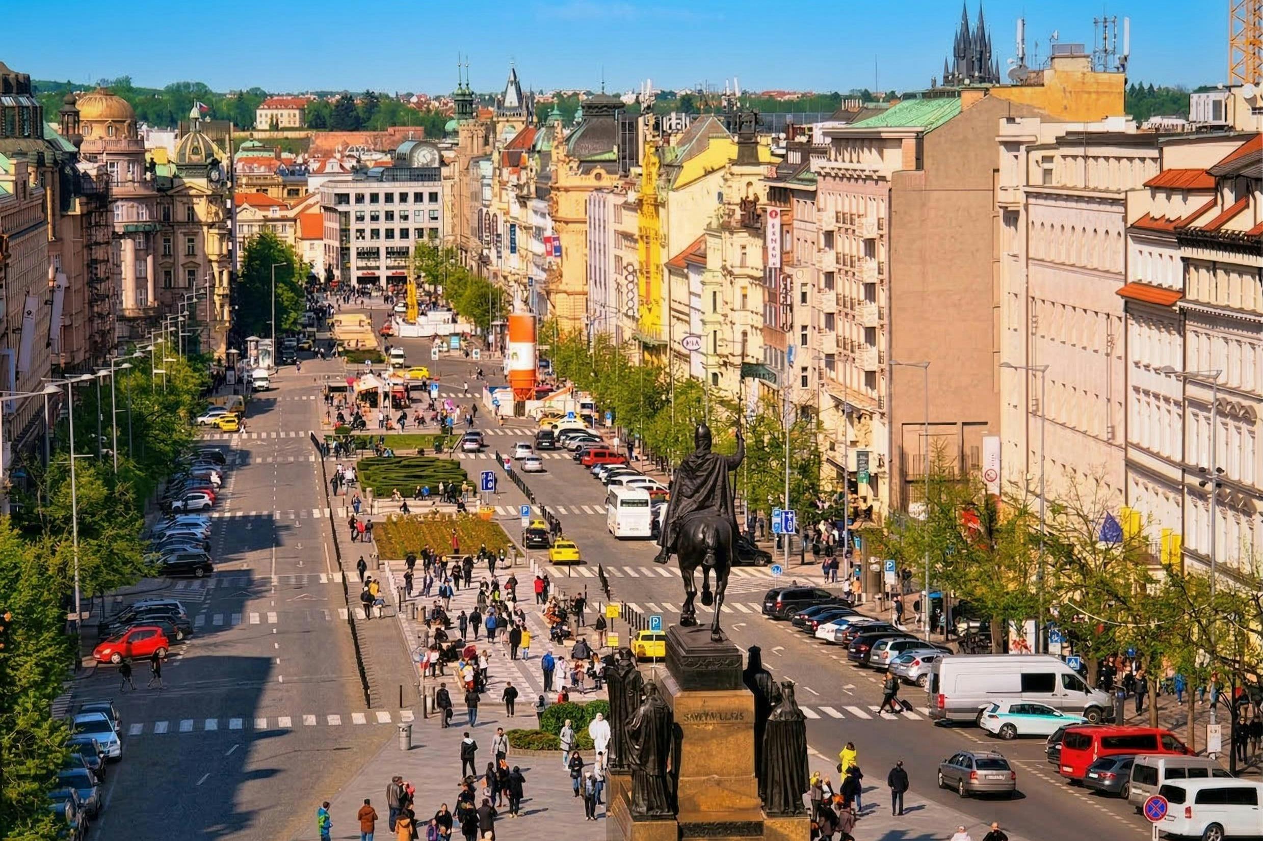 Una bulliciosa plaza de la ciudad con una estatua en primer plano, rodeada de edificios, árboles y gente paseando.