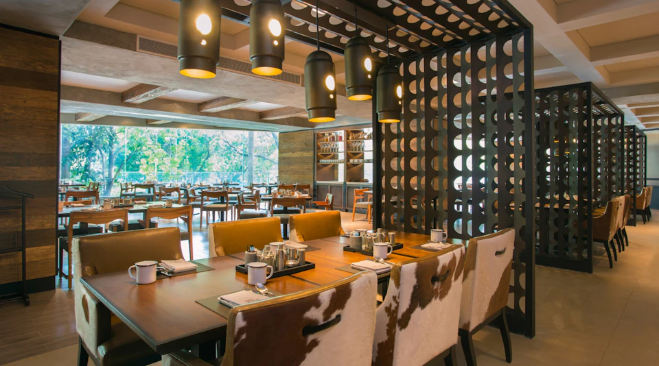 Modern restaurant interior with wooden tables, leather chairs, decorative lighting, and a view of greenery through large windows.