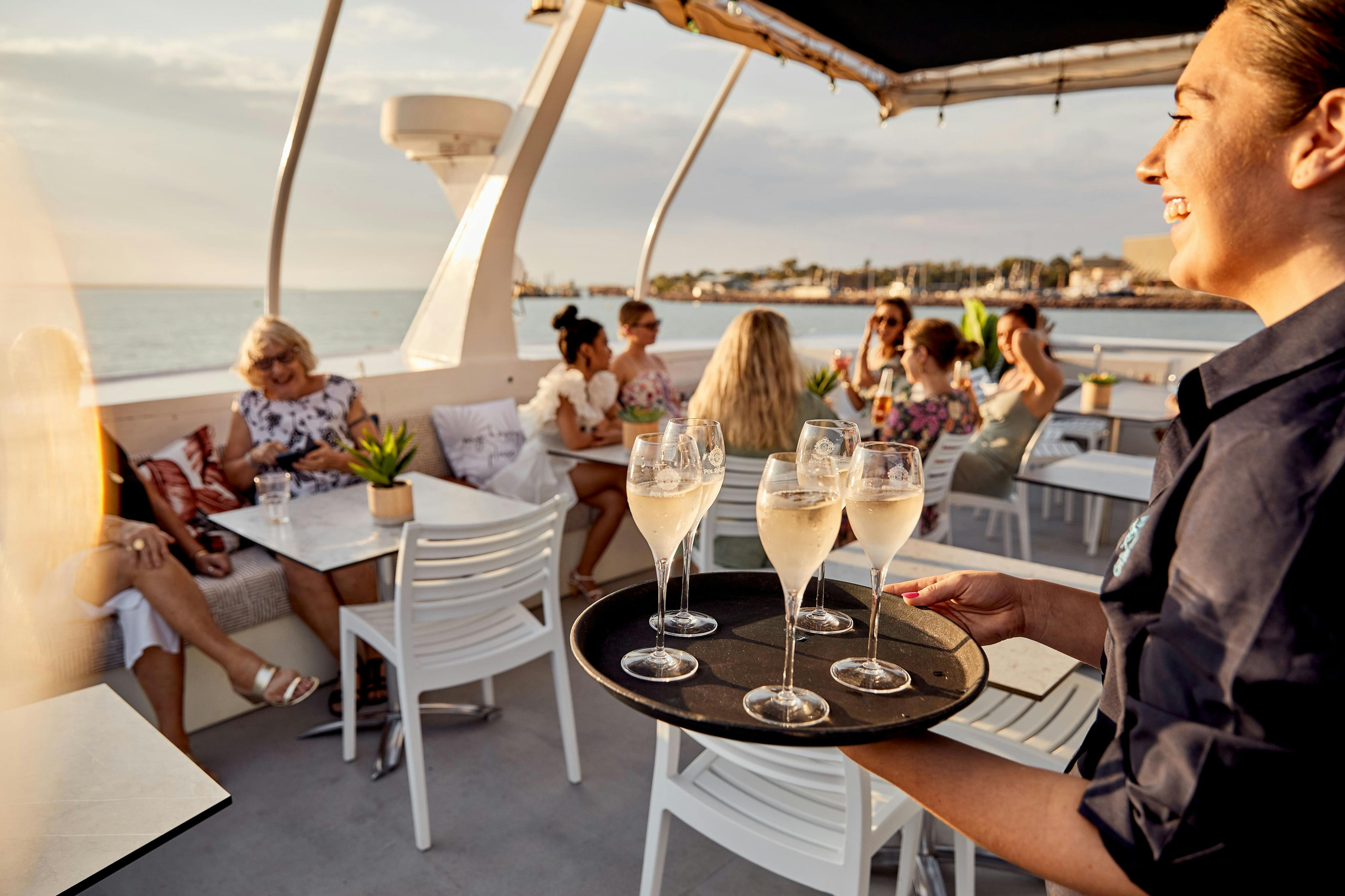 笑顔のウェイトレスがボートデッキでシャンパングラスのトレイを運んでいて、バックグラウンドで人と交流しています。